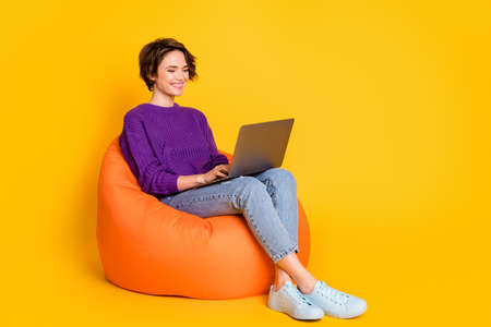 Full length photo portrait of woman working on laptop sitting in orange beanbag chair isolated on vivid yellow colored backgroundの写真素材
