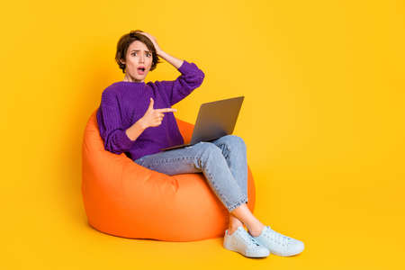 Full length photo portrait of upset girl pointing finger at laptop sitting in orange beanbag isolated on vivid yellow colored backgroundの写真素材
