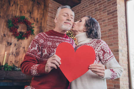 Close-up pphoto portrait couple celebrating winter holidays together keeping red heart postcard wife kissing husbandの写真素材