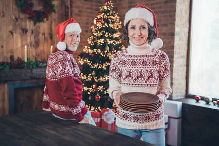 Photo of positive nice pensioner couple set table lady hold pile plates wear ornament sweater hat home indoorsの写真素材