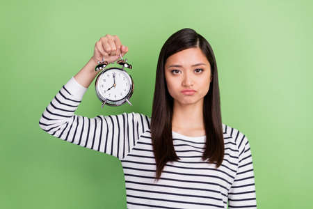 Portrait of attractive depressed tired girl holding in hands clock bedtime wakeup isolated over green color backgroundの写真素材
