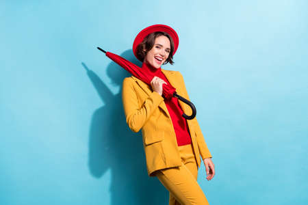 Photo of flirty adorable young lady wear yellow blazer red cap smiling holding parasol isolated blue color backgroundの写真素材