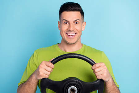 Photo of cheerful young amazed happy man hold hands steering wheel isolated on blue color backgroundの写真素材