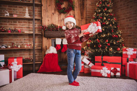 Photo of cheerful glad funny small child girl hold present box wear pullover santa headwear socks indoorsの写真素材