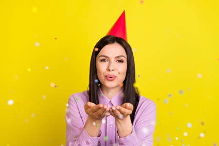 Photo of hooray brown hairdo young lady blow confetti wear birthday hat pink shirt isolated on yellow color backgroundの写真素材