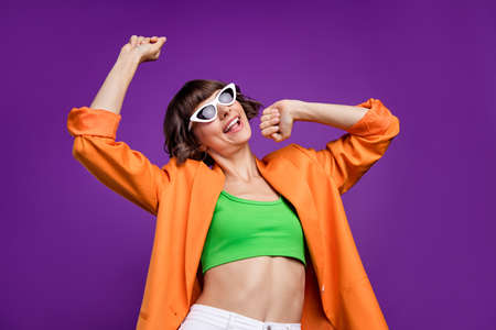 Photo of shiny funky young lady wear orange jacket dark glasses smiling dancing isolated violet...