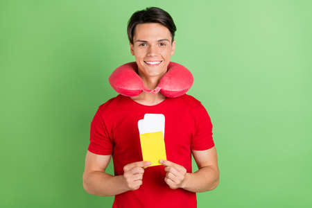 Photo of charming attractive young man wear red t-shirt neck pillow holding tickets isolated green...