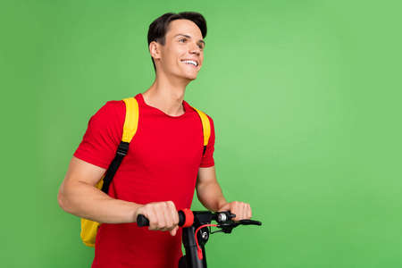 Photo of sweet funky young man wear red t-shirt smiling riding electric scooter looking empty space...