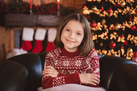 Photo of funny shiny schoolgirl dressed red sweater sitting arms crossed smiling indoors room home houseの写真素材