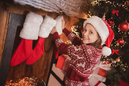 Photo of glad funny joyful small girl decorating house chimney wear ornament sweater santa hat indoorsの写真素材