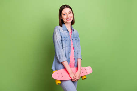 Photo of cool brunette hair young lady hold skate wear jeans jacket isolated on green color backgroundの写真素材