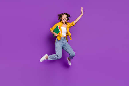 Photo of pretty excited young woman dressed yellow shirt smiling jumping high holding copybooks isolated purple color backgroundの写真素材