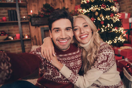 Photo of funky couple do selfie hug wear ugly new year jumpers indoors near tree on x-mas eveの写真素材