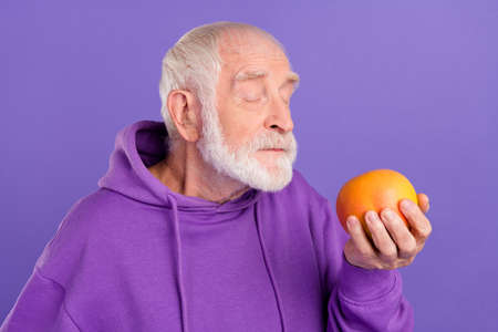 Portrait of attractive dreamy grey-haired man smelling tasty grapefruit isolated over bright violet purple color backgroundの写真素材