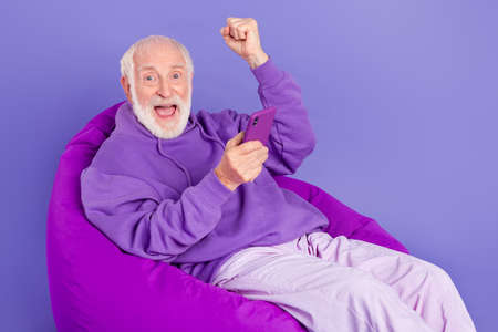 Portrait of elderly retired trendy cheerful lucky grey-haired man sitting in chair rejoice isolated...
