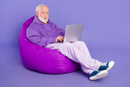 Portrait of elderly retired trendy cheery man pensioner sitting in bag writing email isolated over bright violet purple color backgroundの写真素材