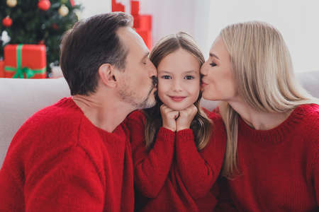 Portrait of attractive cheerful family wife husband parenthood sitting on cozy sofa mom dad kissing schoolgirl december at home indoorsの写真素材