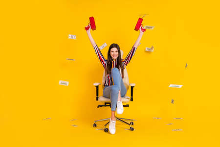 Full body photo of young girl happy smile sit chair shoot movey waste rich dollars air isolated over yellow color backgroundの写真素材