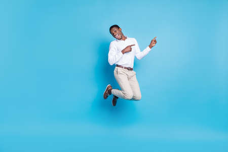 Photo of positive funky dark skin man wear white shirt smiling jumping high pointing empty space isolated blue color backgroundの写真素材