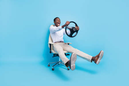 Photo of sweet funky dark skin man wear white shirt smiling sitting chair holding steering wheel isolated blue color backgroundの写真素材