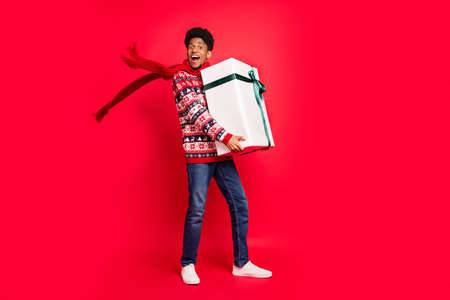 Full length photo of young brunet guy hold present wear holiday scarf sweater jeans shoes isolated on red backgroundの写真素材