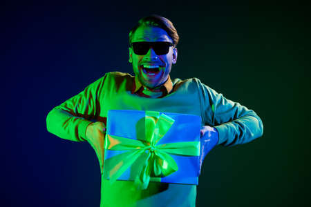Photo portrait of man wearing sunglass keeping wrapped present box on party isolated dark color backgroundの写真素材
