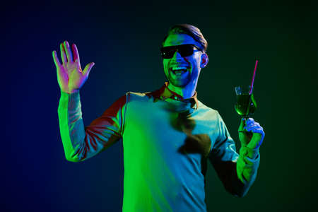 Photo of charming young positive man hold hand cocktail wave hello club smile isolated on neon backgroundの写真素材