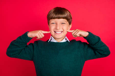 Photo of childish fooling schoolboy dressed green sweater fingers cheeks smiling isolated red color backgroundの写真素材