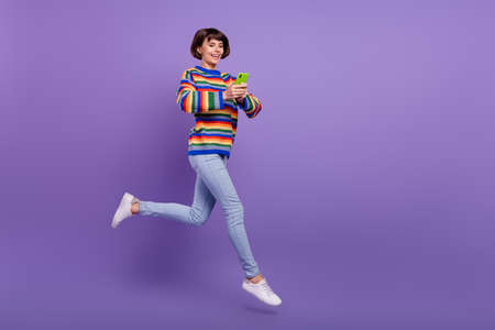 Photo of cheerful lady jump hold phone go texting wear striped sweater jeans sneakers isolated...