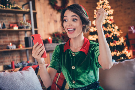 Photo of young shocked amazed positive santa helper woman hold phone winner indoors inside house homeの写真素材