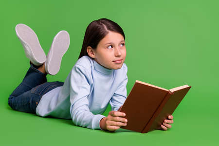Photo of pretty cute schoolgirl wear blue turtleneck lying floor reading book looking empty space smiling isolated green color backgroundの写真素材