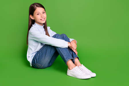 Photo of adorable attractive school girl dressed blue turtleneck smiling sitting floor empty space isolated green color backgroundの写真素材