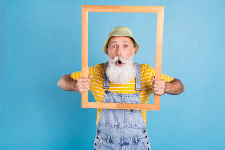 Photo of aged man amazed shocked surprised hold wooden frame photographing isolated over blue color backgroundの写真素材