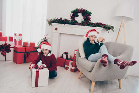 Photo of two curious funny kids prepare unpack x-mas present boxes wear jumper hat in decorated home indoorsの写真素材