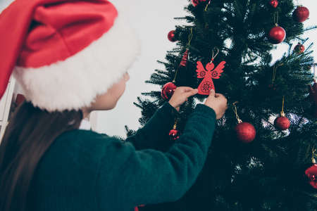 Rear back view photo of small lady hang toys evergreen fir tree wear jumper hat in decorated home indoorsの写真素材