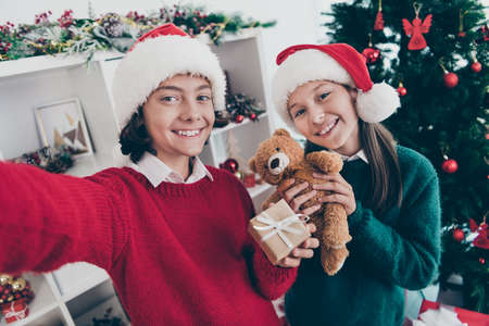 Photo of funny social media blogger kids make self portrait hold presents wear x-mas hat jumper in decorated home indoorsの写真素材