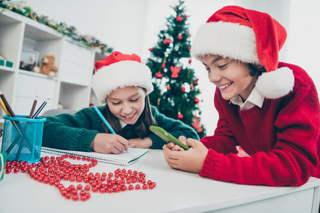 Photo of funny children sit table write santa letter wish gifts wear x-mas hat jumper in decorated home indoorsの写真素材