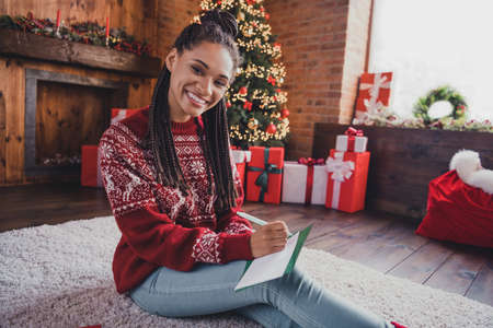 Portrait of attractive cheerful girl sitting on carpet writing mail letter wishlist newyear tradition at decorated home indoorsの写真素材