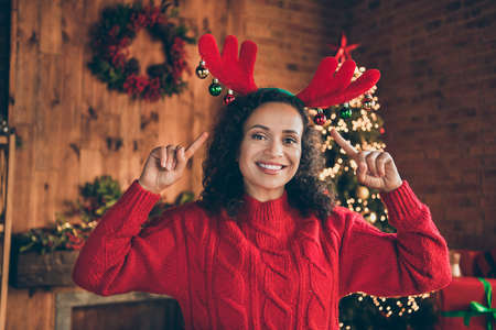 Photo of pretty shiny mature woman wear red sweater smiling pointing fingers new year headband indoors house home roomの写真素材