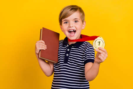 Photo portrait little boy wearing golden medal prize smartest student keeping book isolated vibrant yellow color backgroundの写真素材