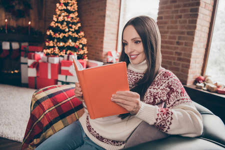 Portrait of attractive focused cheerful woman sitting on cosy divan reading book fairy at brick loft...