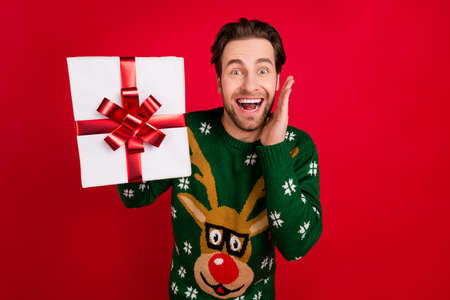 Photo of happy cheerful smiling excited amazed man wear ugly sweater hold gift box isolated on red color backgroundの写真素材