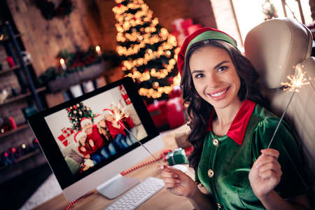 Photo of positive happy woman santa helper talk camera online hold xmas sparklers indoors inside...