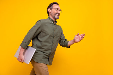 Profile side photo of mature man happy positive smile hold laptop go meeting isolated over yellow color backgroundの写真素材