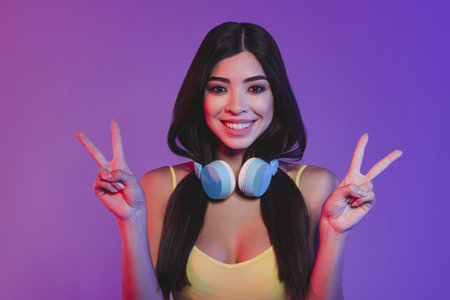 Portrait of attractive cheerful girl listening sound hit showing v-sign isolated over shine purple...