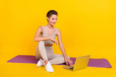 Portrait of attractive thin cheerful girl sitting on mat using laptop lesson isolated over bright...