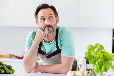 Photo of dreamy inspired thoughtful aged man fist cheek look up wear apron blue t-shirt home kitchen...