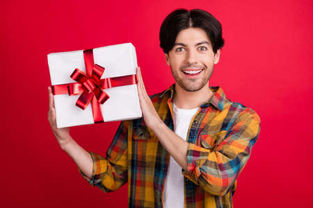 Photo of funny attractive young gentleman wear plaid outfit smiling holding present box isolated red color backgroundの写真素材
