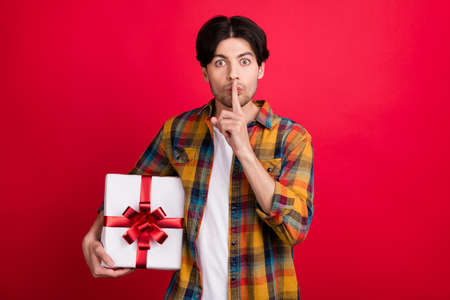 Photo of serious strict young guy dressed checkered clothes asking keep silence about gift isolated red color backgroundの写真素材