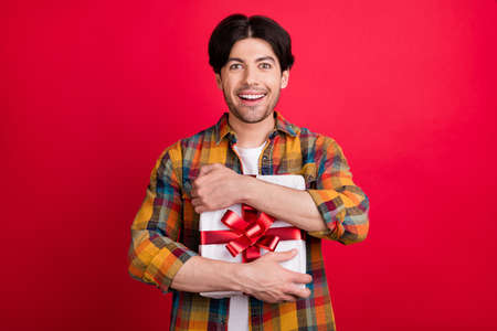 Photo of excited funny young gentleman wear plaid outfit smiling hugging present box isolated red color backgroundの写真素材